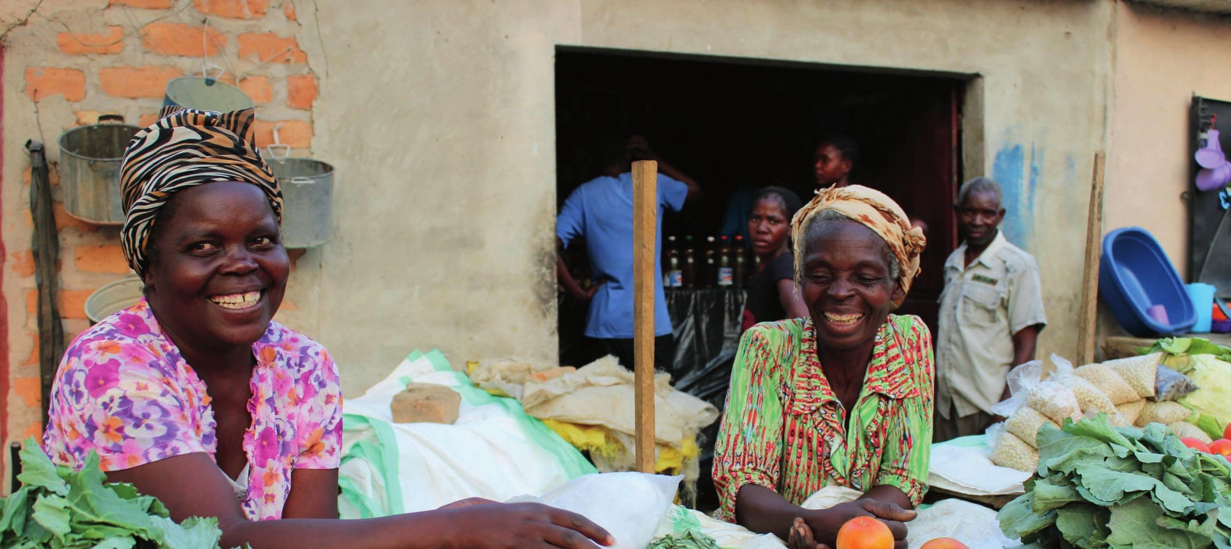 Two smiling women traders at their market stall, with fresh greens and produce in front of them.