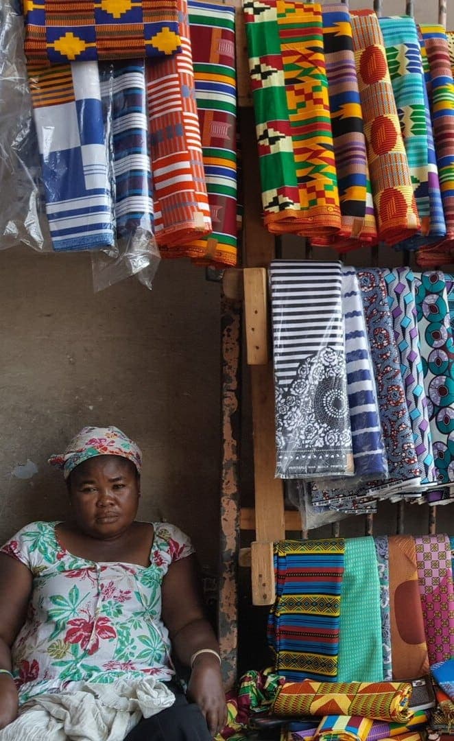 A fabric trader sits beside shelves of kente and wax-print cloth at Makola Market, Accra.