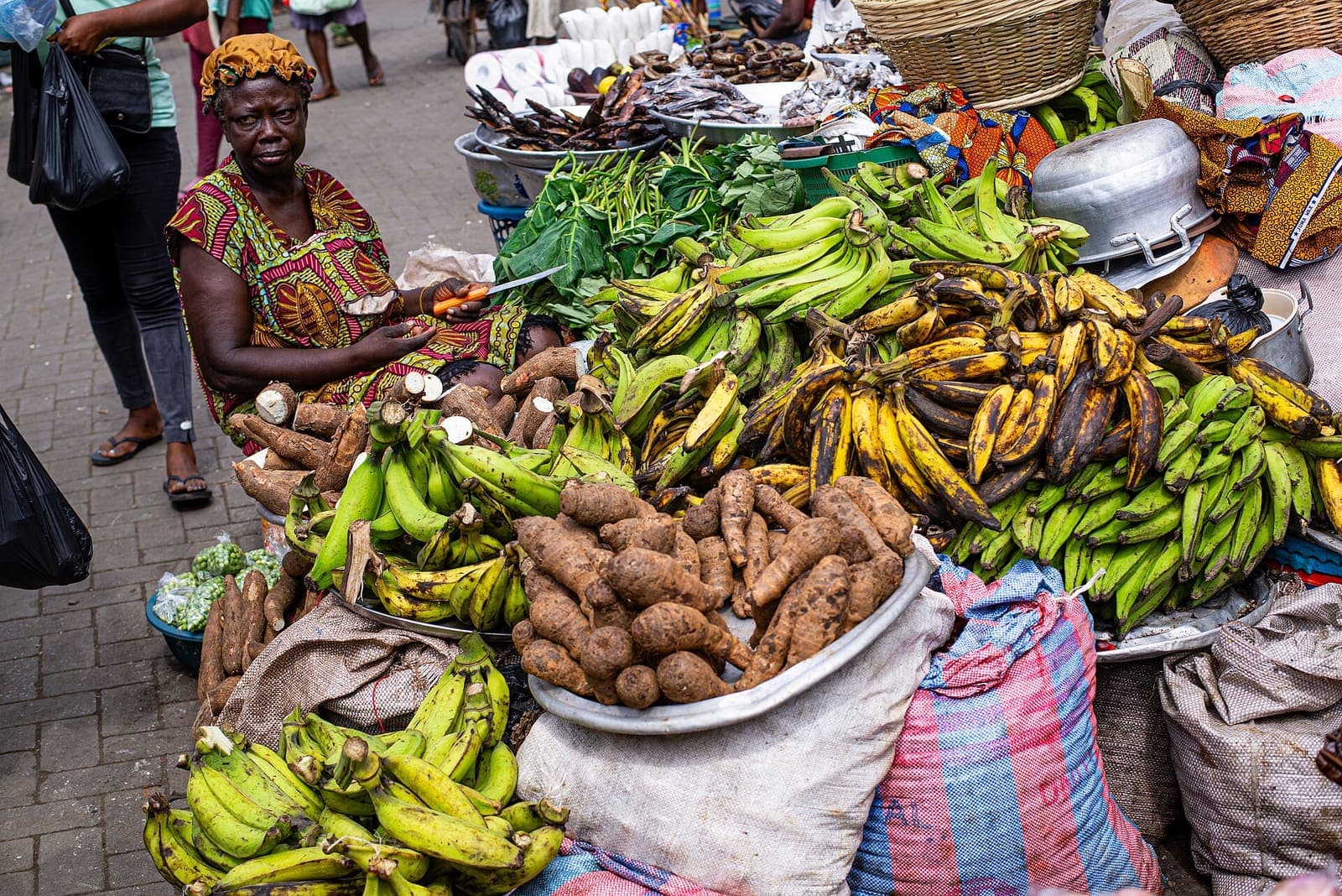 A market trader sits beside her stock of plantain, yam and vegetables.